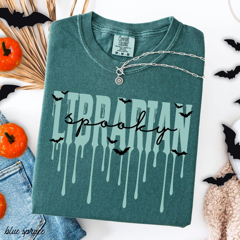 K&ouml;nnte beinhalten: T&uuml;rkisgr&uuml;nes T-Shirt mit den Worten "Librarian Spooky" in einer tropfenden Schriftart, verziert mit Fledermaus-Silhouetten. Eine silberne Halskette mit Sonnenanh&auml;nger liegt auf dem Shirt. Halloween-Dekoration.