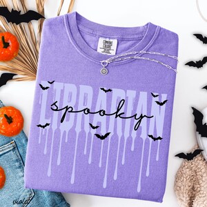 K&ouml;nnte beinhalten: Ein violettes Comfort Colors T-Shirt mit den Worten "LIBRARIAN spooky" in einer tropfenden Schriftart, verziert mit schwarzen Fledermaus-Silhouetten. Eine silberne Halskette mit Sonnenanh&auml;nger befindet sich auf dem Shirt. Orangefarbene K&uuml;rbisse und Papierflederm&auml;use sind im Hintergrund.