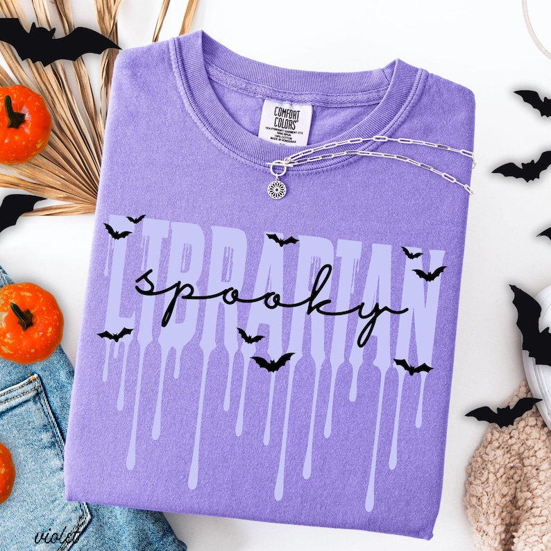 K&ouml;nnte beinhalten: Ein violettes Comfort Colors T-Shirt mit den Worten "LIBRARIAN spooky" in einer tropfenden Schriftart, verziert mit schwarzen Fledermaus-Silhouetten. Eine silberne Halskette mit Sonnenanh&auml;nger befindet sich auf dem Shirt. Orangefarbene K&uuml;rbisse und Papierflederm&auml;use sind im Hintergrund.