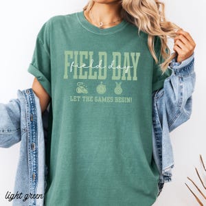 May include: Light green t-shirt with the words "FIELD DAY" in a white and green font. Below the text are three small icons: a shoe, a stopwatch, and a medal. The phrase "LET THE GAMES BEGIN!" is printed below the icons.