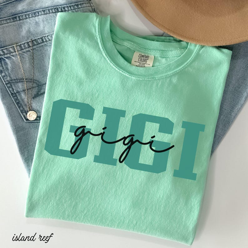 Op de afbeelding: Een lichtgroen t-shirt met de naam "GIGI" gedrukt in turquoise blokletters met een cursief lettertype. Het shirt is gemaakt van een zacht materiaal en wordt getoond met een blauwe jeans en een bruine hoed.