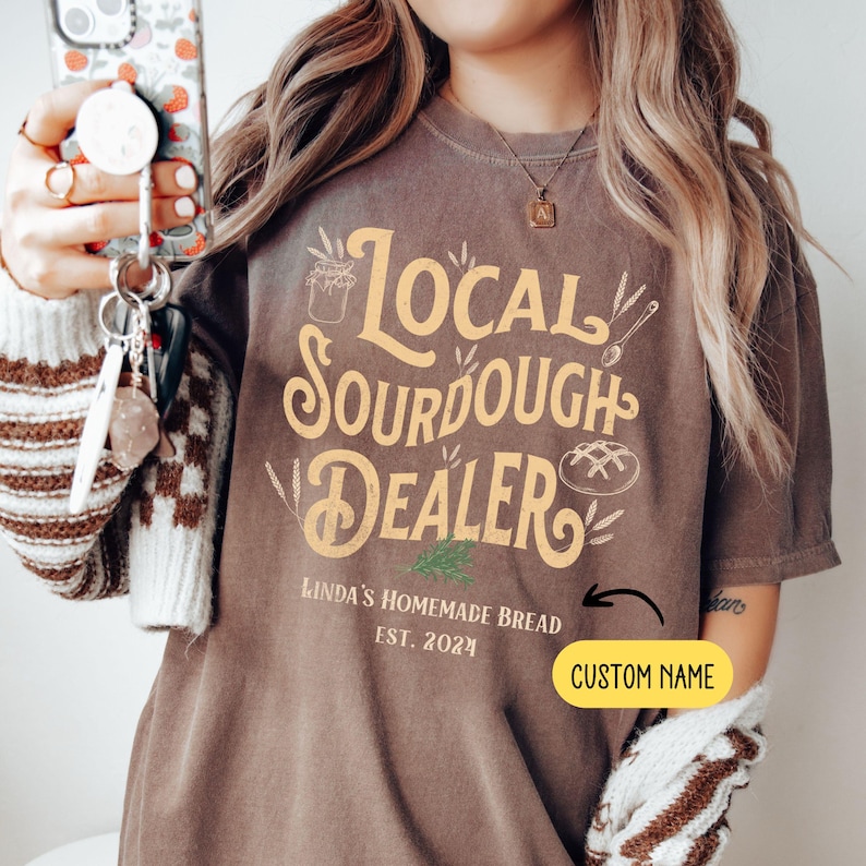 Peut inclure: Un t-shirt marron avec le texte "Local Sourdough Dealer" en jaune. Le texte "Linda's Homemade Bread Est. 2021" est sous le texte principal. Il y a un espace pour un nom personnalis&eacute;.