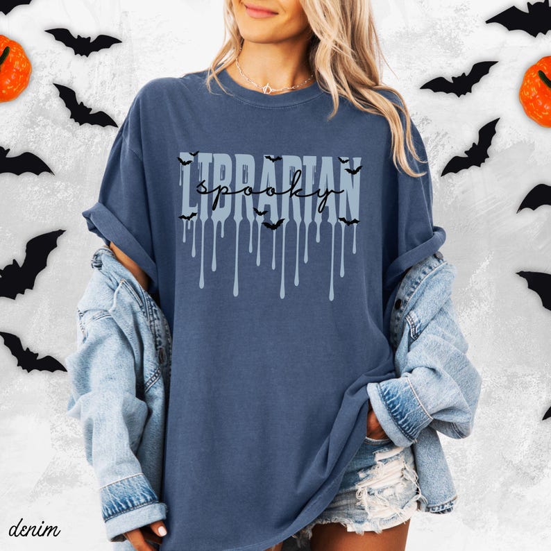 K&ouml;nnte beinhalten: Ein dunkelblaues T-Shirt mit dem Aufdruck "Librarian Spooky" in hellblauer Schrift, Tropfen-Akzenten und Fledermaus-Grafiken. Das Shirt geh&ouml;rt zu einer Halloween-Kollektion.