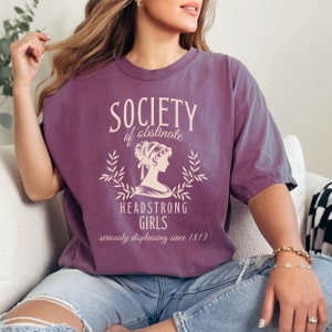 May include: A purple t-shirt with a vintage-style graphic that reads "Society of Obstinate Headstrong Girls seriously displeasing since 1813".