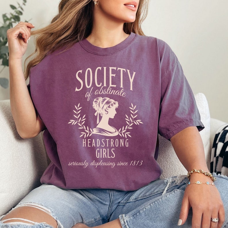 May include: A purple t-shirt with a vintage-style graphic that reads "Society of Obstinate Headstrong Girls seriously displeasing since 1813".