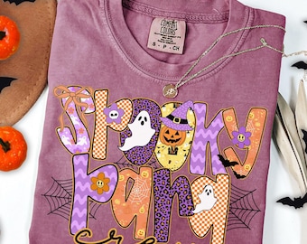 May include: A berry-colored t-shirt featuring the text "SPOOKY PALS crew" in large, patterned letters. The design includes ghosts, a jack-o'-lantern in a witch hat, flowers, spiderwebs, and bats in shades of orange, purple, and white.