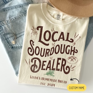 Peut inclure: Un t-shirt beige avec du texte brun qui dit "Local Sourdough Dealer" et "Linda's Homemade Bread Est. 2024". Il y a un dessin d'une miche de pain, d'un bocal et d'&eacute;pis de bl&eacute;. Le texte "Custom Name" est dans une bo&icirc;te jaune.