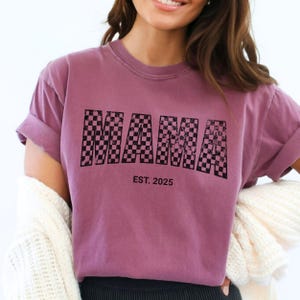 Pu&ograve; includere: T-shirt color malva con la scritta "MAMA" in un motivo a scacchi neri sul davanti. Sotto, il testo "EST. 2025". La maglietta ha maniche corte e una vestibilit&agrave; ampia.