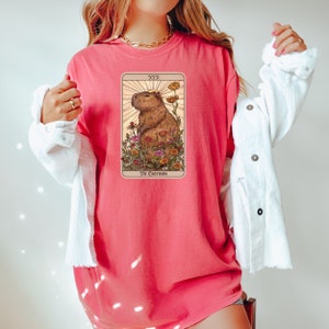 Pode incluir: Uma camiseta rosa com um gr&aacute;fico de uma capivara em um campo de flores. O gr&aacute;fico &eacute; uma carta de tar&ocirc; com o texto "XIX" e "THE CAPYBARA".