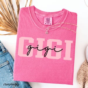 Op de afbeelding: Een roze Comfort Colors t-shirt met de naam "GIGI" gedrukt in een lichtroze blokletter, met "gigi" in een cursief lettertype. Een zilveren ketting met een zonnebloemhanger ligt op het shirt.