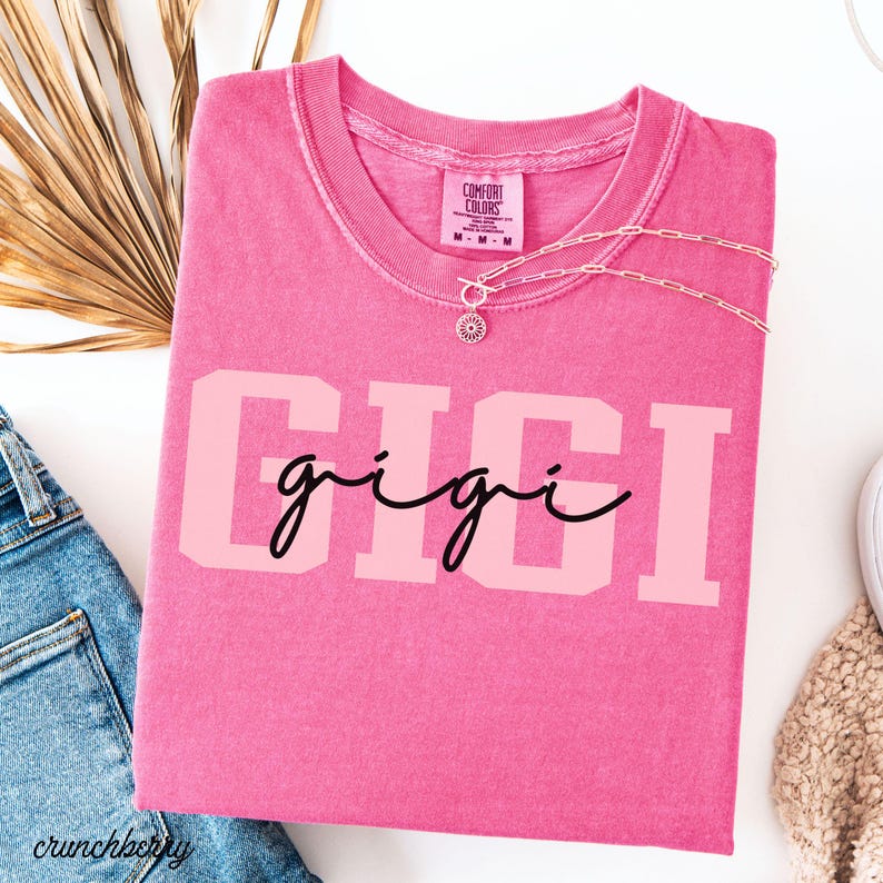 Op de afbeelding: Een roze Comfort Colors t-shirt met de naam "GIGI" gedrukt in een lichtroze blokletter, met "gigi" in een cursief lettertype. Een zilveren ketting met een zonnebloemhanger ligt op het shirt.