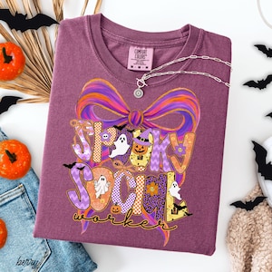 May include: A berry-colored t-shirt featuring a large graphic of a colorful bow and the text "Spooky Social Worker." The design includes illustrations of ghosts, pumpkins, flowers, and spiderwebs in shades of purple, orange, and yellow.