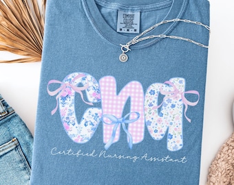 May include: A blue Comfort Colors t-shirt with the letters "CNA" in a floral and gingham pattern, accented with bows. The text "Certified Nursing Assistant" is below the letters. A silver necklace with a pendant is on the shirt.
