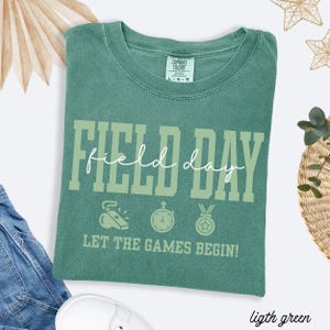 May include: Light green t-shirt with the words "FIELD DAY" in large, light green block letters. Below the text are three small illustrations of a whistle, stopwatch, and medal. The phrase "LET THE GAMES BEGIN!" is printed below the illustrations.