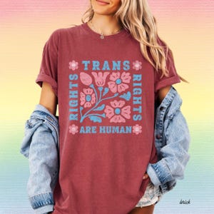 May include: Brick-colored t-shirt with the text "TRANS RIGHTS ARE HUMAN" in a square design. The design includes pink flowers and blue accents. The shirt has rolled-up sleeves and a casual fit.