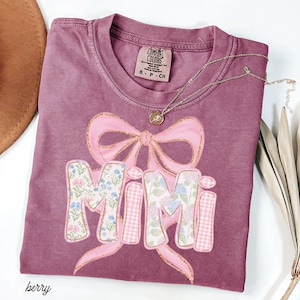 May include: Berry-colored t-shirt with a pink bow and the word "MIMI" in a floral and gingham pattern. A gold necklace with a pendant is draped across the shirt. The shirt is labeled "Comfort Colors" and is size S-CH.