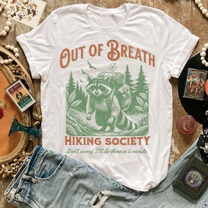 Pode incluir: Camiseta branca com um guaxinim verde usando uma mochila e caminhando pela floresta. O texto "Out of Breath Hiking Society" est&aacute; impresso na camiseta, junto com a frase "N&atilde;o se preocupe, estarei l&aacute; em um minuto".