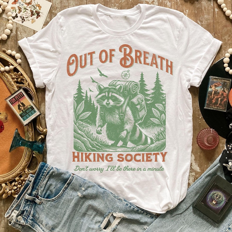 Pode incluir: Camiseta branca com um guaxinim verde usando uma mochila e caminhando pela floresta. O texto "Out of Breath Hiking Society" est&aacute; impresso na camiseta, junto com a frase "N&atilde;o se preocupe, estarei l&aacute; em um minuto".