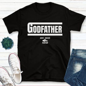 May include: Black t-shirt with white text that reads "Godfather Est. 2025 Noah" with a small graphic of two hands clasped together.