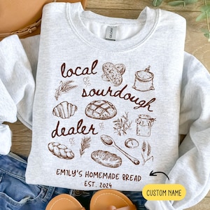 May include: A gray sweatshirt with a brown graphic design of bread, flour, and a spoon. The text on the graphic reads "local sourdough dealer" and "Emily's Homemade Bread Est. 2024". There is a space for a custom name.