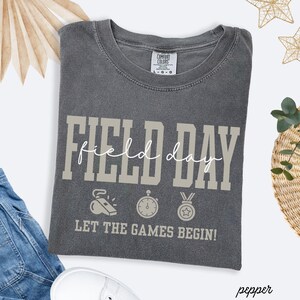 May include: A gray Comfort Colors t-shirt with the words "FIELD DAY" in a block font and "field day" in a script font. Below are illustrations of a whistle, stopwatch, and medal. The text "LET THE GAMES BEGIN!" is below the illustrations.