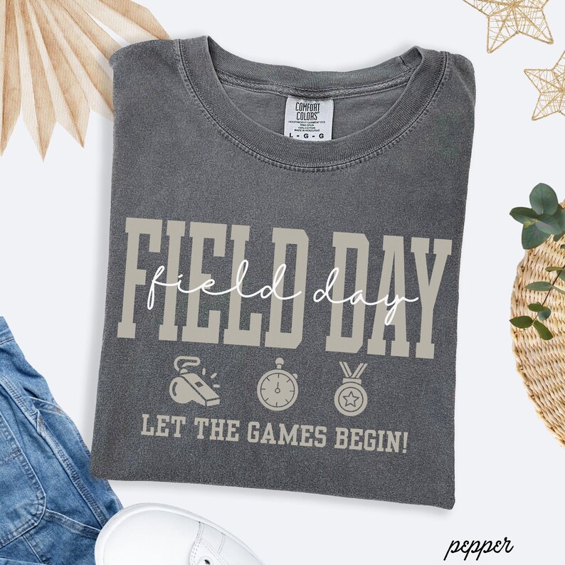May include: A gray Comfort Colors t-shirt with the words "FIELD DAY" in a block font and "field day" in a script font. Below are illustrations of a whistle, stopwatch, and medal. The text "LET THE GAMES BEGIN!" is below the illustrations.