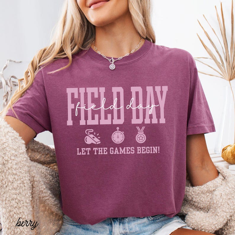 May include: Berry-colored t-shirt with the words "FIELD DAY" in large, light pink letters. Below the text are three small icons: a whistle, a stopwatch, and a medal. The phrase "LET THE GAMES BEGIN!" is printed below the icons.