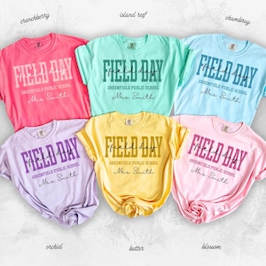May include: Six short-sleeved t-shirts in various colors, including pink, blue, and yellow. Each shirt features the words "FIELD DAY" in large, bold letters, along with the text "GREENFIELD PUBLIC SCHOOL Mrs. Smith."