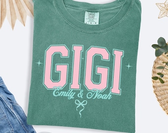 Könnte beinhalten: Hellgrünes T-Shirt mit dem Namen "GIGI" in großen rosa Buchstaben mit hellgrüner Umrandung. Darunter stehen die Worte "Emily & Noah" und eine dekorative Schleife. Das Shirt ist aus weichem Material.