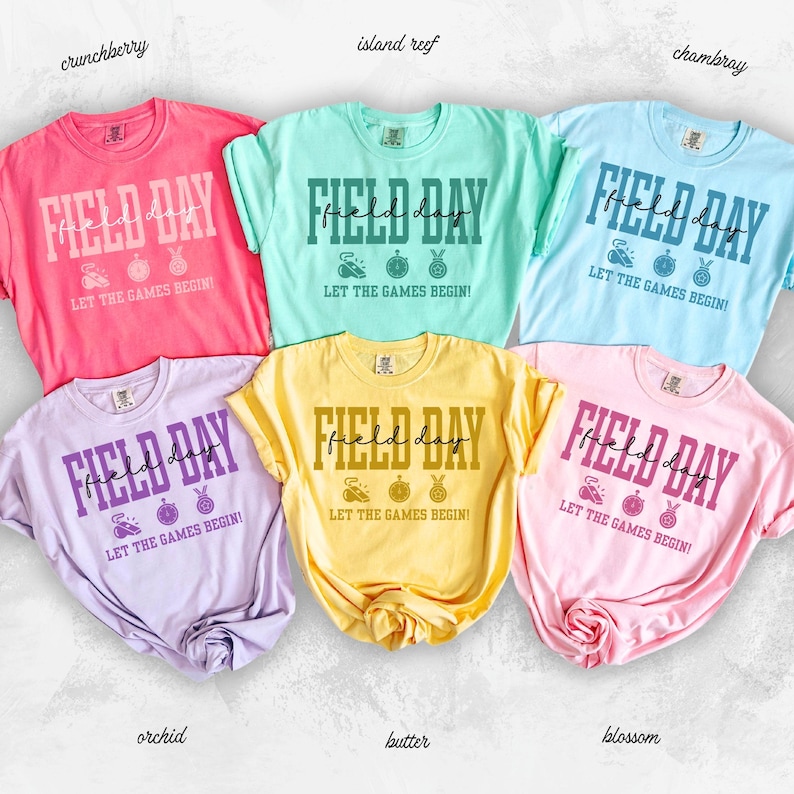 May include: A collection of six short-sleeved t-shirts in various colors, including pink, blue, yellow, and purple. Each shirt features the words "FIELD DAY" in a bold font, along with sports-themed graphics and the phrase "LET THE GAMES BEGIN!"