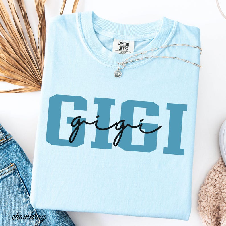 Op de afbeelding: Een lichtblauw Comfort Colors T-shirt met de naam "GIGI" in een tweekleurig ontwerp op de voorkant. Het woord "GIGI" is vetgedrukt, met het woord "gigi" in cursief. Een zilveren ketting met een hanger ligt op het T-shirt.