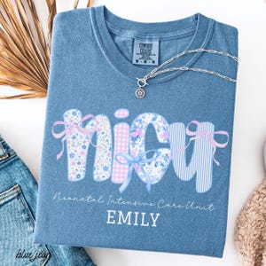 May include: Blue Comfort Colors t-shirt with the word "nicu" in a floral, gingham, and striped design, accented with bows. The text "Neonatal Intensive Care Unit" and the name "EMILY" are below the word. A silver necklace with a pendant is on the shirt.