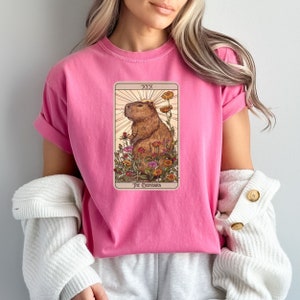Pode incluir: Camiseta rosa com um gr&aacute;fico de uma capivara cercada de flores. O gr&aacute;fico est&aacute; no estilo de uma carta de tar&ocirc; e diz "XIX The Capybara".
