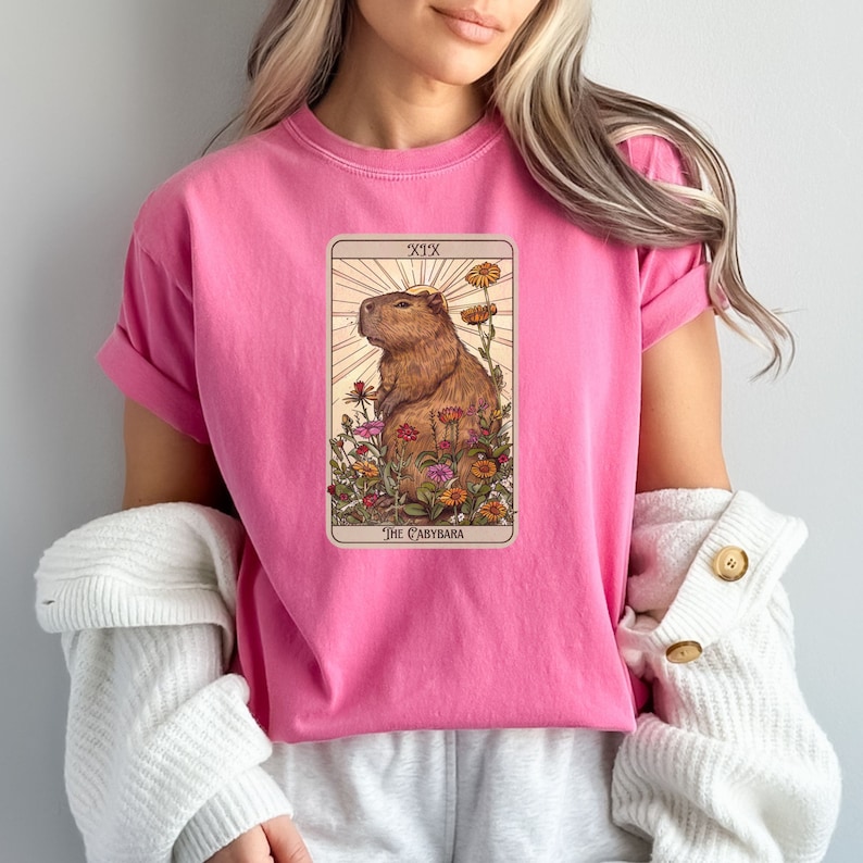 Pode incluir: Camiseta rosa com um gr&aacute;fico de uma capivara cercada de flores. O gr&aacute;fico est&aacute; no estilo de uma carta de tar&ocirc; e diz "XIX The Capybara".