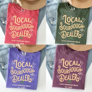 Peut inclure: Quatre t-shirts de couleurs diff&eacute;rentes avec le texte "Local Sourdough Dealer" dans une police de caract&egrave;res de style vintage. Le texte est en jaune avec un contour brun. Les chemises sont rouge, verte, violette et marron.
