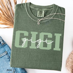 Op de afbeelding: Mosgroen t-shirt met de naam "GIGI" in grote, lichtgroene blokletters, met "gigi" in een cursief lettertype over de blokletters. Een zilveren ketting met een zonnebloemhanger ligt op het shirt. Het shirt is opgevouwen.