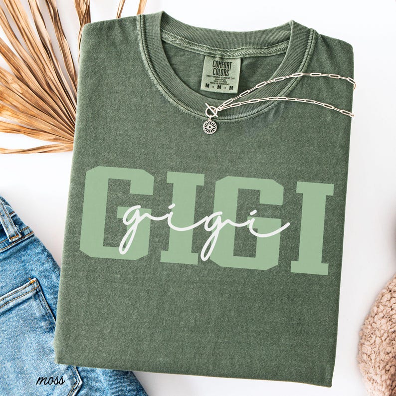 Op de afbeelding: Mosgroen t-shirt met de naam "GIGI" in grote, lichtgroene blokletters, met "gigi" in een cursief lettertype over de blokletters. Een zilveren ketting met een zonnebloemhanger ligt op het shirt. Het shirt is opgevouwen.