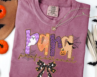 May include: A berry-colored t-shirt with a graphic print that reads "para professional" in large, patterned letters featuring ghosts, pumpkins, spiderwebs, and leopard print. Below the text is a black bow with a ghost and pumpkin pattern.