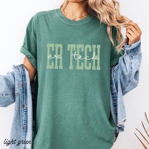 May include: A light green t-shirt with the text "ER TECH" in a block letter style. The words are in a lighter shade of green. The shirt is a solid color and has rolled-up sleeves.
