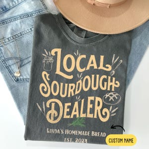 Peut inclure: Un t-shirt gris avec le texte "Local Sourdough Dealer" en jaune. Le texte est entour&eacute; d'&eacute;pis de bl&eacute; et d'un pot de levain. Le texte "Linda's Homemade Bread Est. 2024" est sous le texte principal. Il y a un espace pour un nom personnalis&eacute;.