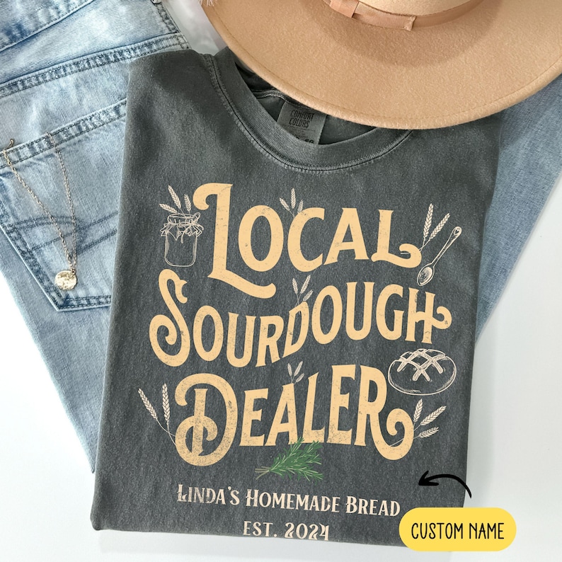 Peut inclure: Un t-shirt gris avec le texte "Local Sourdough Dealer" en jaune. Le texte est entour&eacute; d'&eacute;pis de bl&eacute; et d'un pot de levain. Le texte "Linda's Homemade Bread Est. 2024" est sous le texte principal. Il y a un espace pour un nom personnalis&eacute;.