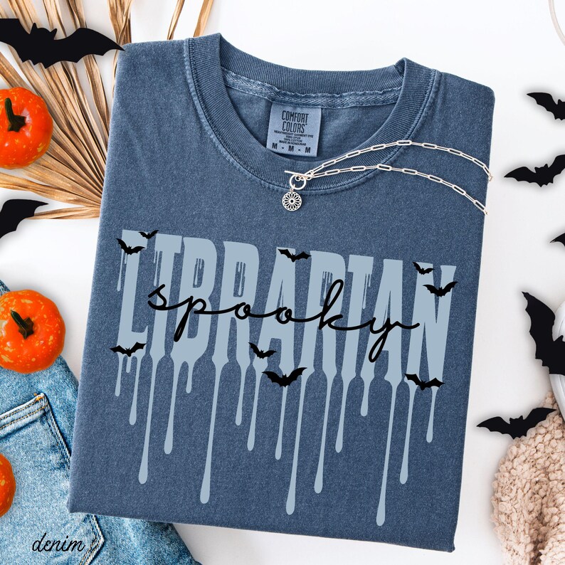 K&ouml;nnte beinhalten: Ein jeansblaues T-Shirt mit den Worten "LIBRARIAN spooky" in einer gruseligen Schriftart. Das Shirt hat eine silberne Halskette mit einem Sonnenanh&auml;nger. Schwarze Fledermaus-Ausschnitte und orangefarbene K&uuml;rbisse schm&uuml;cken den Hintergrund.