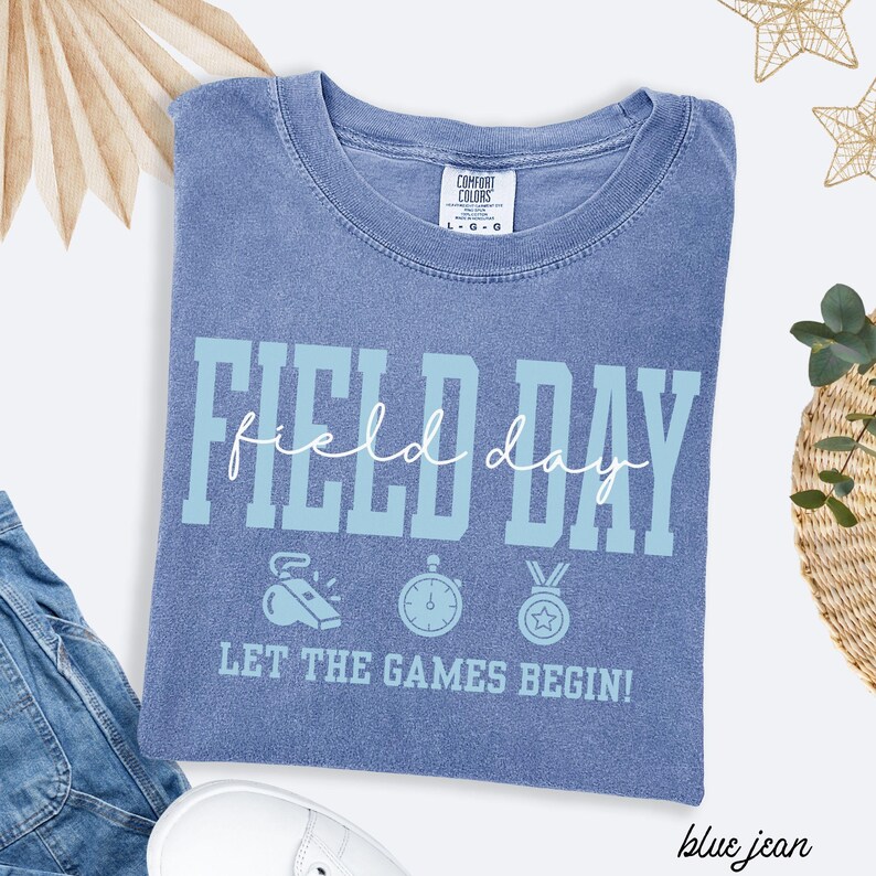 May include: Blue jean-colored t-shirt with the words "FIELD DAY" in light blue block letters. The shirt also features the text "LET THE GAMES BEGIN!" and sports-themed graphics. The shirt is made of a soft, comfortable material.