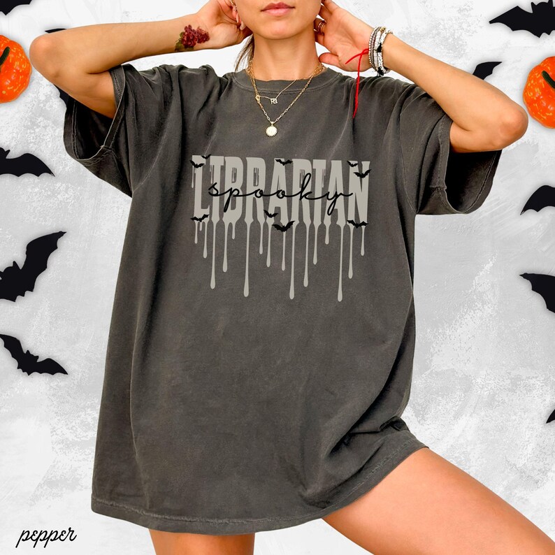 K&ouml;nnte beinhalten: Dunkelgraues T-Shirt mit dem Schriftzug "Librarian Spooky" in tropfender Schrift und Fledermaus-Silhouetten. Ein lockeres T-Shirt, ideal f&uuml;r Halloween oder die gruselige Jahreszeit.