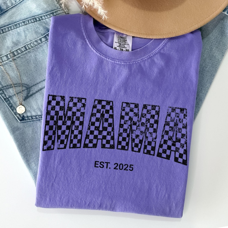 Pu&ograve; includere: Una maglietta viola con la scritta "MAMA" in un motivo a scacchi bianco e nero. Il testo "EST. 2025" &egrave; sotto. La maglietta &egrave; piegata sopra un paio di jeans blu e un cappello marrone.