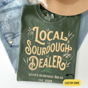 May include: Green t-shirt with the text "Local Sourdough Dealer" in a vintage style font. The text is in a light yellow color. Below the text is a smaller text that says "Linda's Homemade Bread Est. 2024" and a space for a custom name.
