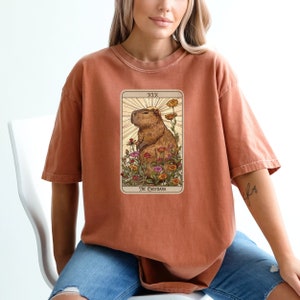 Pode incluir: Uma camiseta cor ferrugem com um gr&aacute;fico de uma capivara em um campo de flores. O gr&aacute;fico &eacute; uma carta de tar&ocirc; com o texto "XIX The Capybara".