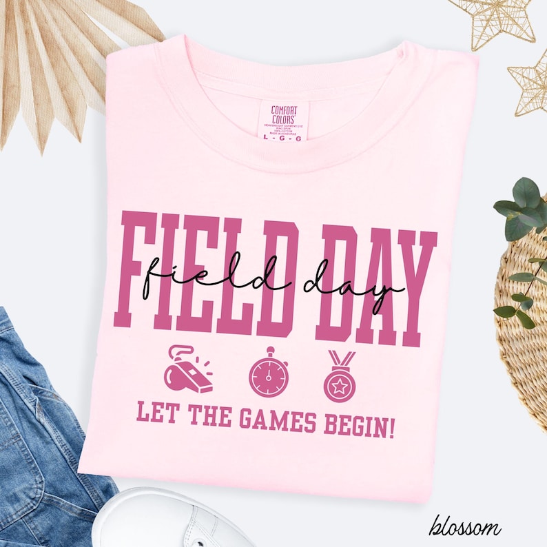May include: A light pink t-shirt with the words "FIELD DAY" in a bold, dark pink font. Below the text are three small icons: a whistle, a stopwatch, and a medal. The phrase "LET THE GAMES BEGIN!" is printed below the icons.