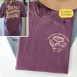 Pu&ograve; includere: Una maglietta viola con il testo "Local Sourdough Dealer" sul retro e un disegno di diversi pani con il testo "Emma's Rustic Homemade Bread Est. 2021" sul davanti.