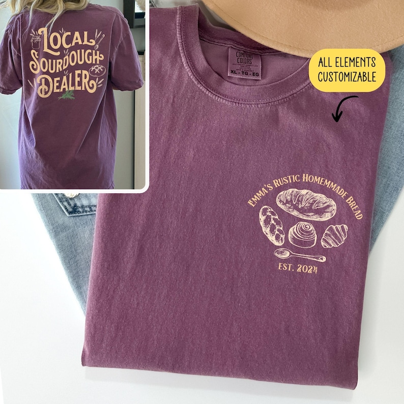 Pu&ograve; includere: Una maglietta viola con il testo "Local Sourdough Dealer" sul retro e un disegno di diversi pani con il testo "Emma's Rustic Homemade Bread Est. 2021" sul davanti.